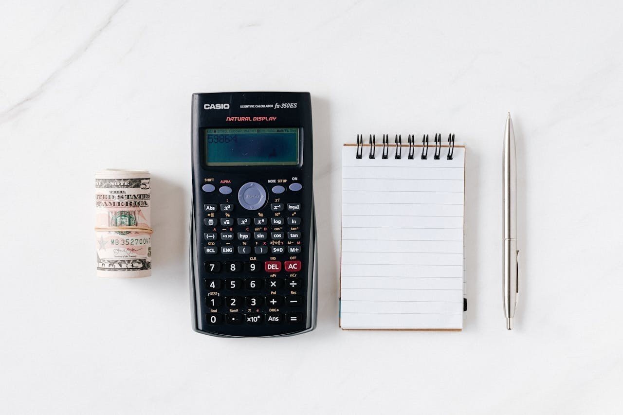 Dollar bills, calculator, notes, and a pen on a white surface.